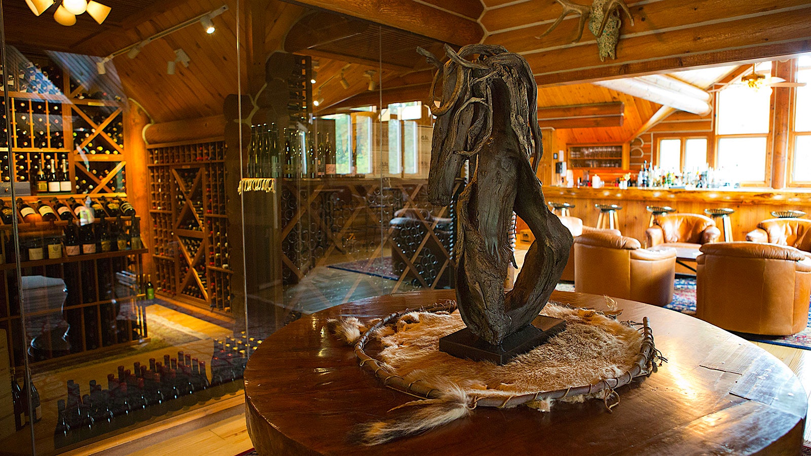 Leather chairs and a bar at Triple Creek Ranch, near a round wooden table, a rustic wood sculpture, a deer trophy hanging on the wooden wall and a cellar of wine bottles